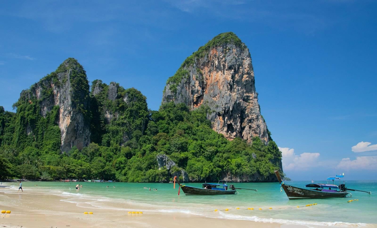 A beach with boats and a large rock formation in the background with Railay Beach in the background